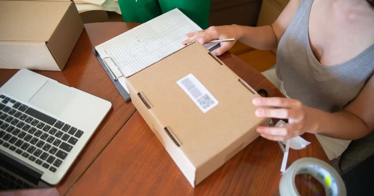 Người phụ nữ đóng gói đơn hàng vào hộp giấy bên cạnh máy tính xách tay